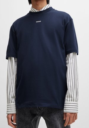Osoba nosząca granatowy t-shirt Hugo nałożony na białą koszulę z długim rękawem w czarne pionowe paski oraz ciemnoszare dżinsy.