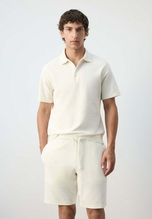 Joven de pie con una camiseta tipo polo de manga corta color blanco roto y pantalones cortos a juego con cordón, sobre un fondo liso.