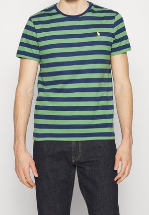 Homme portant une chemise à manches courtes rayée vert et bleu marine et un jean bleu foncé, debout les bras détendus le long du corps.