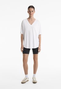 Chemise blanche à manches courtes en V, coupe décontractée, associée à un short de vélo noir. Le modèle porte des baskets beiges avec des chaussettes blanches. Design simple.