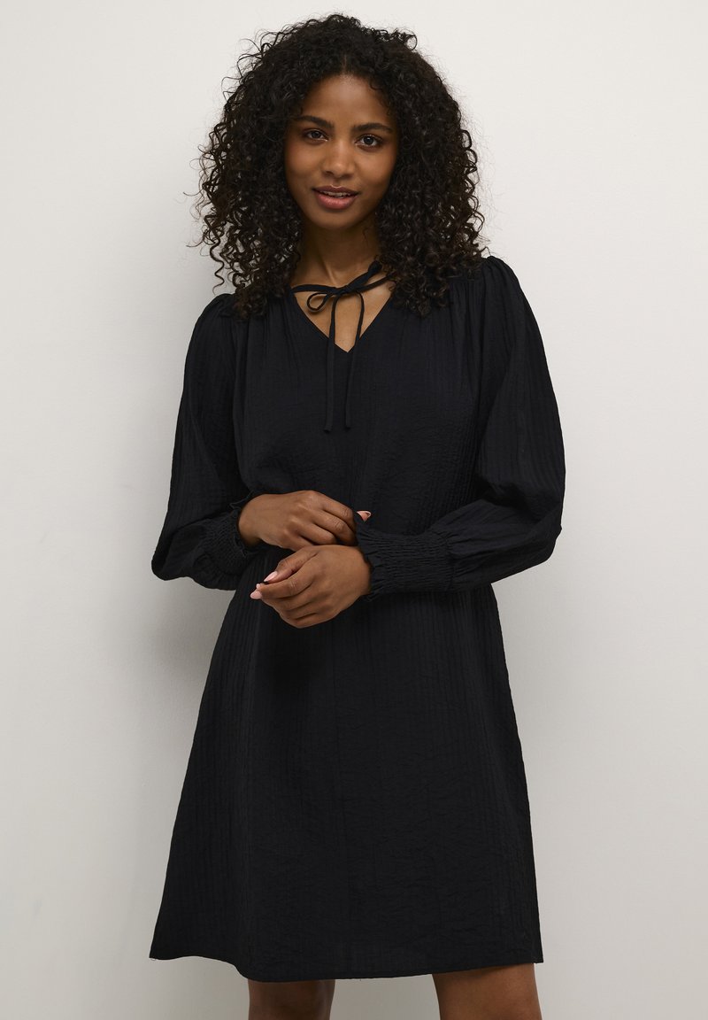 Robe noire en tissu texturé, décolleté en V et détails à nouer ; manches longues bouffantes avec des poignets élastiques ; longueur au genou.