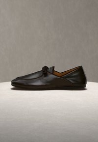 Μαύρο δερμάτινο παπούτσι τύπου slip-on με μικρή λεπτομέρεια κόμπου στην κορυφή, εμφανιζόμενο από το πλάι σε φωτεινή επιφάνεια με ουδέτερο φόντο.