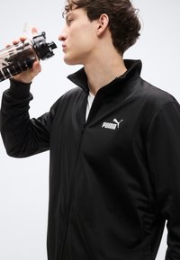 Μαύρο μπουφάν με φερμουάρ και ψηλό γιακά, κατασκευασμένο από λείο υλικό. Διαθέτει λευκό λογότυπο Puma στο αριστερό στήθος και κομψό, εφαρμοστό σχέδιο.