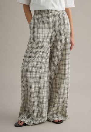 Persona que lleva pantalones beige de tiro alto y pernera ancha con patrón de cuadrícula negra, camisa blanca de manga corta y sandalias negras de punta abierta.