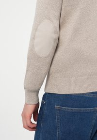 Jasno beżowy sweter z teksturowanym splotem i łatkami na łokciach z gładkiego materiału, dopasowanymi mankietami oraz prostym krojem. Noszony z niebieskimi dżinsami.