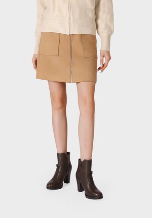 Donna che indossa una minigonna in suede color cammello con tasche frontali e zip, maglione di lana color crema e stivaletti marrone scuro con tacco.