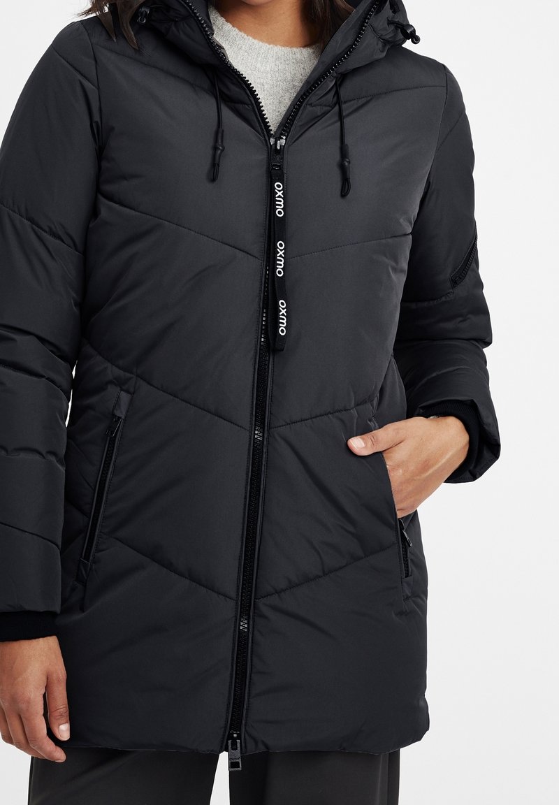 Svart puffjacka med dragkedja fram, huva och dragskor. Har ett diagonalt kviltat mönster och sidofickor för praktisk användning.