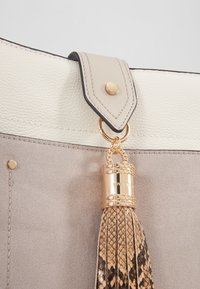 Handväska med en design i krämfärgad och ljusgrå läder, med guldfärgad hårdvara, en dekorativ frans och en slät textur.