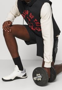 Felpa senza maniche nera con scritte rosse, camicia a maniche lunghe crema sotto, pantaloni corti neri, scarpe da ginnastica bianche e manubrio in mano.