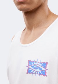 Gros plan d'une personne portant un débardeur blanc avec un logo en forme de soleil bleu et rose contenant la lettre « S » sur la poitrine.