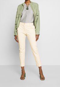 Person som bär ljusbeige slim-fit byxor, grön öppen jacka, randig blus med tofsar och gröna sandaler med hög klack och remmar.