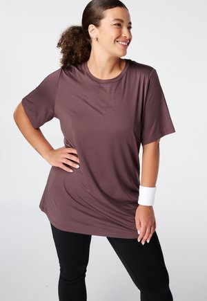 Vrouw lacht en poseert in een losse bruine T-shirt, zwarte leggings en een witte polsband tegen een effen lichte achtergrond.