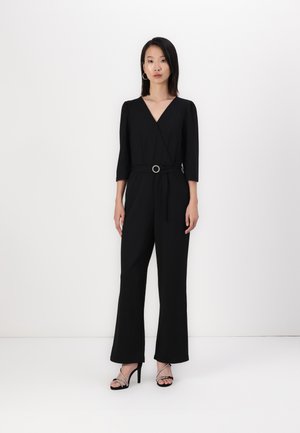 Black jumpsuit with a wrap design, three-quarter sleeves, and a contrasting belt with a circular buckle. Worn with strappy black heels.