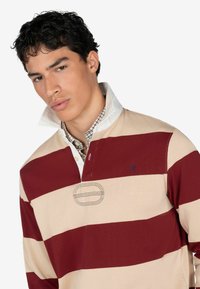 Maglietta da rugby a maniche lunghe con strisce alternate bordeaux e beige, colletto con bottoni e con uno strato interno bianco, e dettaglio del logo ricamato.