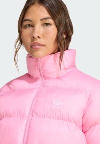 Jonge vrouw met lang, golvend haar draagt een hoogsluitende roze pufferjack met een wit Adidas-logo. Zachte achtergrondtinten.