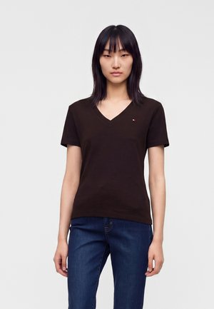 Kvinde med glat sort hår iført en sort V-hals kortærmet skjorte og blå jeans, der står mod en ensfarvet lys baggrund.