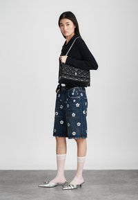 Modella in un maglione nero a maglia, pantaloni shorts di denim con ricami floreali, che tiene in mano una borsa nera trapuntata, indossando mule argentate e calze bianche trasparenti.
