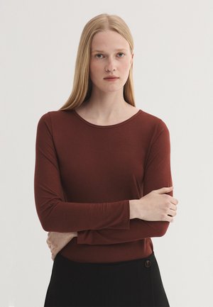 Junge Frau mit langen blonden Haaren, die ein braunes Langarmshirt und schwarze Hosen trägt, steht mit verschränkten Armen vor einem schlichten Hintergrund.