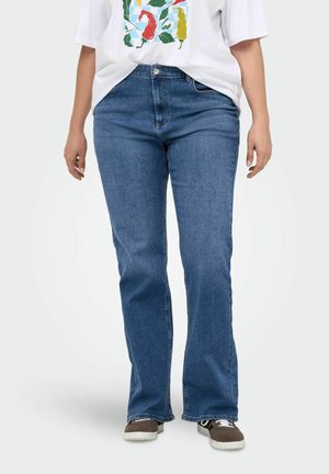 Person iført blå straight-leg jeans, en hvid t-shirt med farverigt design og brune sneakers stående på hvid baggrund.