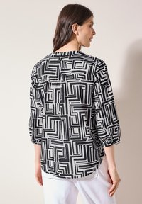 Blusa de patrón en blanco y negro con mangas de tres cuartos, que presenta un diseño geométrico y un dobladillo ligeramente curvado. Tejido ligero.