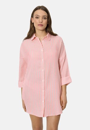 Mujer con un vestido camisa suelto, abotonado, con rayas verticales rosas y blancas y mangas remangadas, de pie contra un fondo liso.