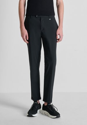 Man draagt strakke, zwarte, op maat gemaakte broek, zwart mouwloos topje en zwarte sneakers met witte zolen, staand tegen een effen grijze achtergrond.