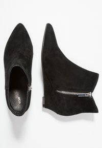 Bottines à talons compensés en suède noir avec un bout pointu, fermeture éclair sur le côté et texture lisse, présentant un design élégant et moderne.