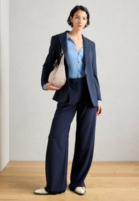 Marineblau gestreifter Blazer über einem hellblauen gerippten Oberteil, kombiniert mit weiten marineblauen Hosen und einer beigen Tasche. Die Schuhe sind spitz mit weißen Akzenten.
