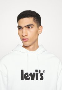 Biela mikina s kapucňou a čiernym logom "Levi's". Má nastaviteľnú kapucňu a rebrované manžety, vyrobená z mäkkej, textúrovanej tkaniny. Jednoduchý dizajn.