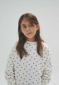 Mikina v off-white barvě s černými puntíky, dlouhé rukávy, žebrovaný kulatý výstřih a jemná textura látky. Jednoduché pozadí.