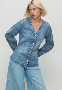 Világoskék farmerdzseki V-nyakkivágással, hosszú ujjakkal és gombos záródással. Testhezálló kialakítással és finom, függőleges varrási részletekkel rendelkezik.