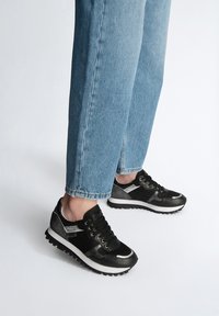 Scarpe da atletica nere e argentate con tomaie in rete, dettagli testurizzati, punta rotonda e suole in gomma bianca, abbinate a jeans azzurri chiari.