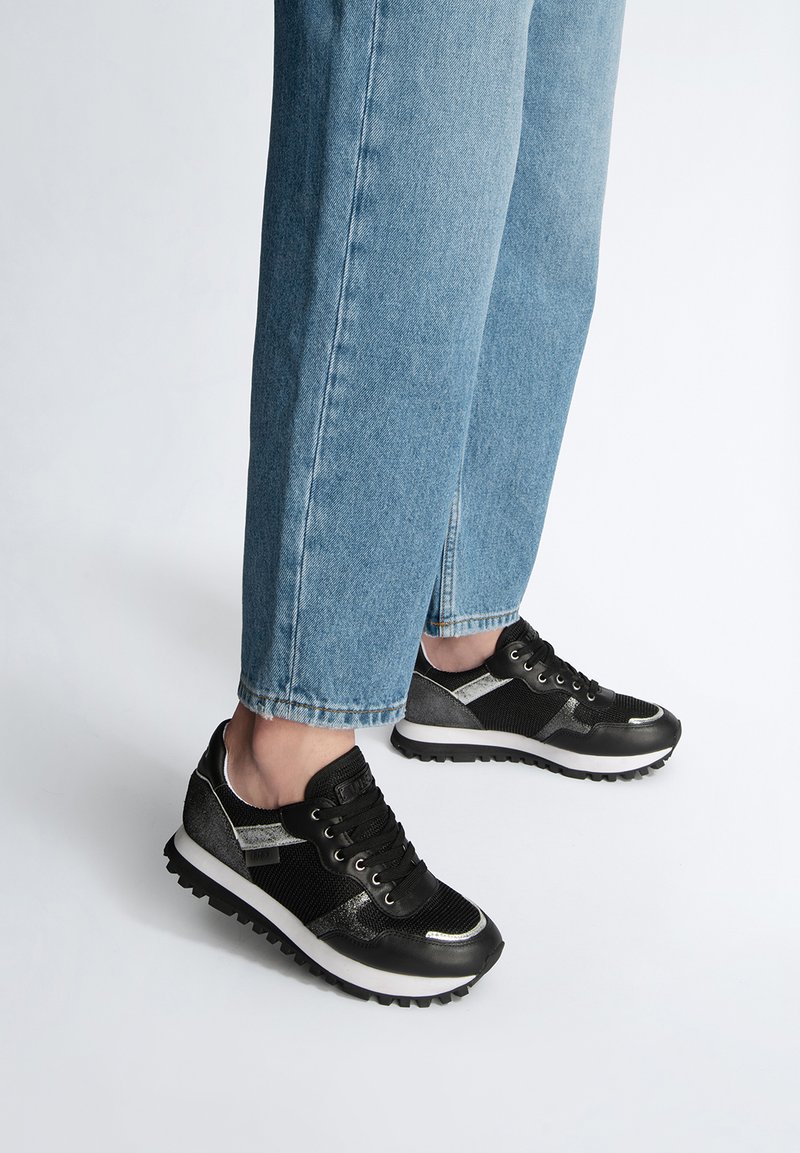 Scarpe da atletica nere e argentate con tomaie in rete, dettagli testurizzati, punta rotonda e suole in gomma bianca, abbinate a jeans azzurri chiari.