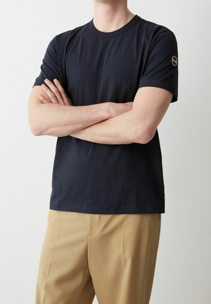 Mann trägt ein schlichtes schwarzes T-Shirt mit kurzen Ärmeln und einem kleinen runden Logo auf dem linken Ärmel sowie beige Hosen, die Arme verschränkt.