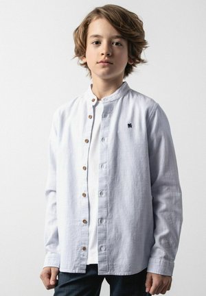 Chłopiec o średniej długości brązowych włosach, ubrany w białą koszulę na guziki na białej koszulce, stoi na tle jednolitego jasnego tła.