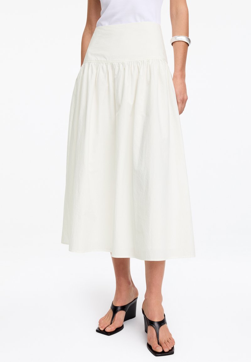 Femme portant une jupe midi blanche froncée, des sandales à talons noires, un haut blanc et un bracelet argenté au bras sur un fond blanc uni.