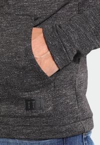 Hand i fickan på en mörkgråmelerad tröja med en "ITD"-patch, person klädd i blå jeans.