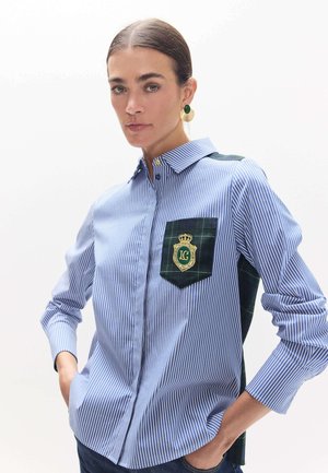 Mujer con el pelo peinado hacia atrás que lleva una camisa de rayas azules y blancas con un bolsillo de cuadros oscuro y un emblema verde dorado, con las manos en los bolsillos.