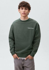 Felpa verde realizzata in morbido tessuto, con collo tondo, polsini a costine e logo "Outdoor Sports" in bianco. Indossata su jeans grigi sbiaditi.