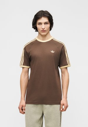 Νέος άνδρας με μεσαίου μήκους σκούρα μαλλιά φοράει καφέ μπλουζάκι Adidas με μπεζ περιγράμματα και ρίγες, σε συνδυασμό με ανοιχτόχρωμο παντελόνι, στέκεται ακίνητος.