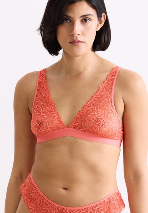 Woman with short dark hair wearing coral lace bralette and matching lace underwear, standing against a plain, light background.