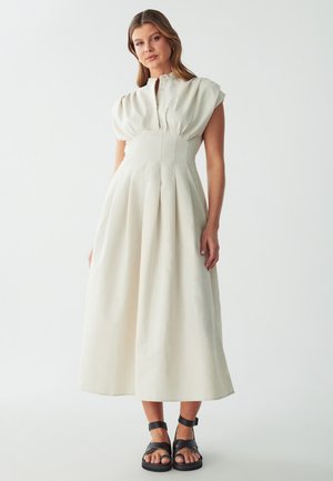 Cream-colored midi dress with pleated skirt, short puff sleeves, and a buttoned neckline. Worn with black strappy sandals.