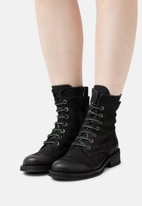 Bottines en cuir noir avec lacets verts, bout rond, finition texturée et petit talon empilé, dotées d'une fermeture éclair latérale pour un enfilage facile.