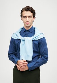 Mėlyna medvilninė marškinėlių su sagomis ir šviesiai mėlynas megztinis, užmestas ant kaklo; modelis demonstruoja neutralų išraišką.