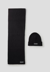Ensemble écharpe et bonnet en tricot noir, côtelé. L'écharpe est longue et rectangulaire ; le bonnet a un revers replié. Les deux présentent une étiquette de logo.