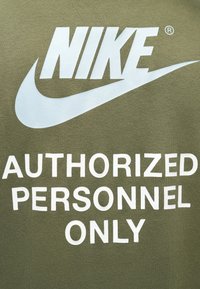 Olivroheline sügisjakk, mis on valmistatud puuvillast, suurte valgete Nike logo ja paksude tähtedega tekstiga "AINULT VOLITATUD ISIKUD".