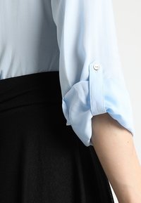 Blusa azzurro chiaro con maniche arrotolate e dettagli con bottoni, abbinata a una gonna nera, che mostra una texture liscia e un design semplice.