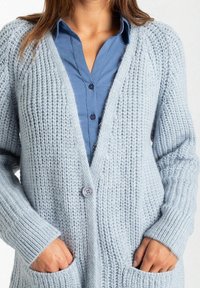 Cardigan lavorato a maglia azzurro chiaro con scollo a V, maniche a costine, tasche frontali e chiusura con un bottone. Tessuto morbido e motivo a maglia grossa.