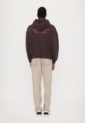 Brauner Hoodie mit dem Text "COMMUNITY OFFICER" in Pink, kombiniert mit hellen, weitem Hosen und Schuhen mit schwarzen Akzenten.