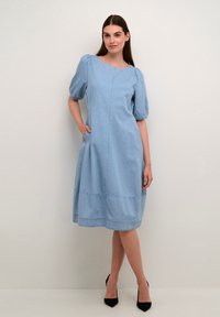 Gai zila džinsa kleita līdz ceļgaliem, ar pūkainām piedurknēm, sānu kabatām un šuvju detaļu vidū. Valkātas melnas augstpapēžu kurpes.